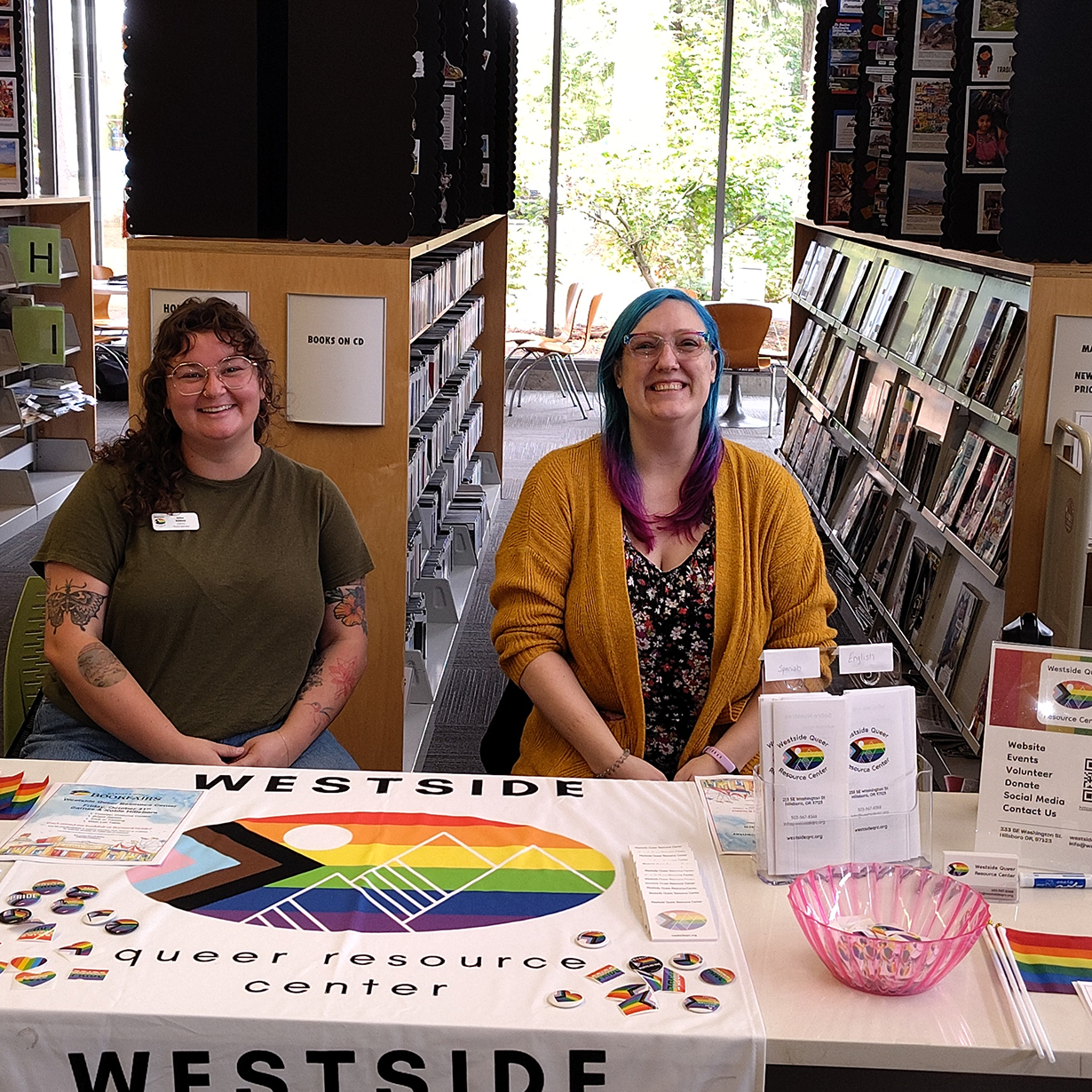 Two smiling women sitting behind a Westside Queer Resource Center table displaying flags and pins inside a library.