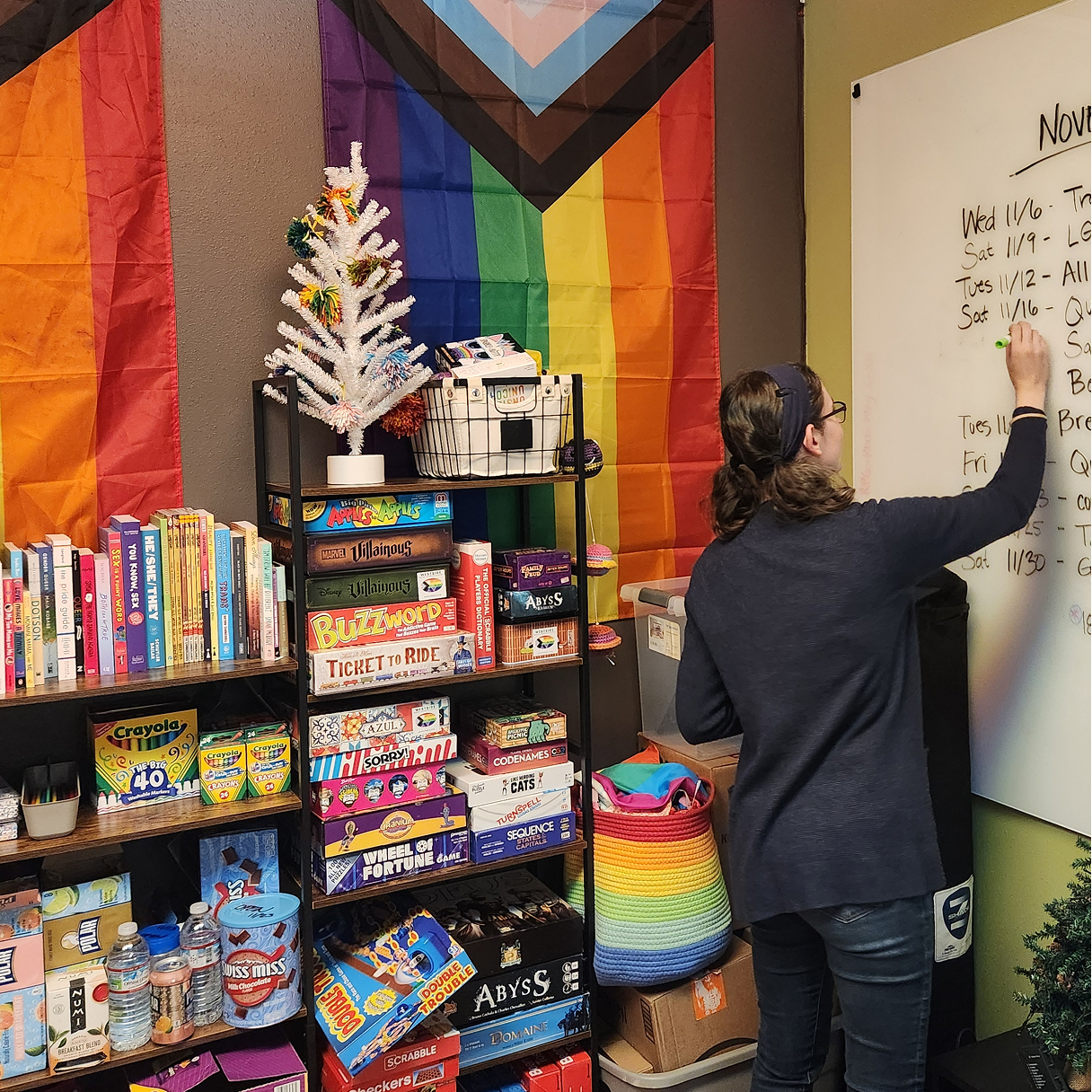 A person writes on a whiteboard next to shelves stocked with board games and books, backed by a Pride flag.